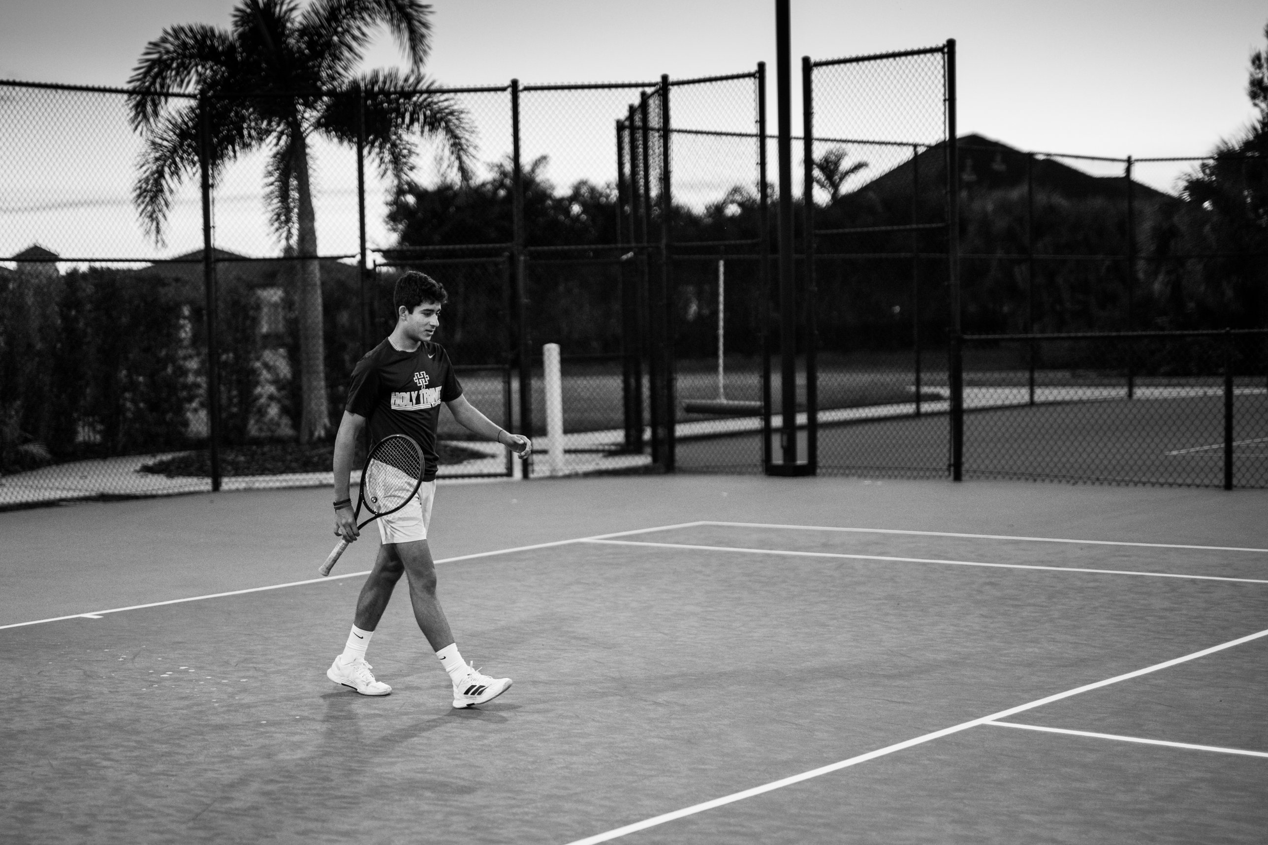 Shaan walking across the court in his Holy Trinity shirt at dusk, palm tree and fence behind