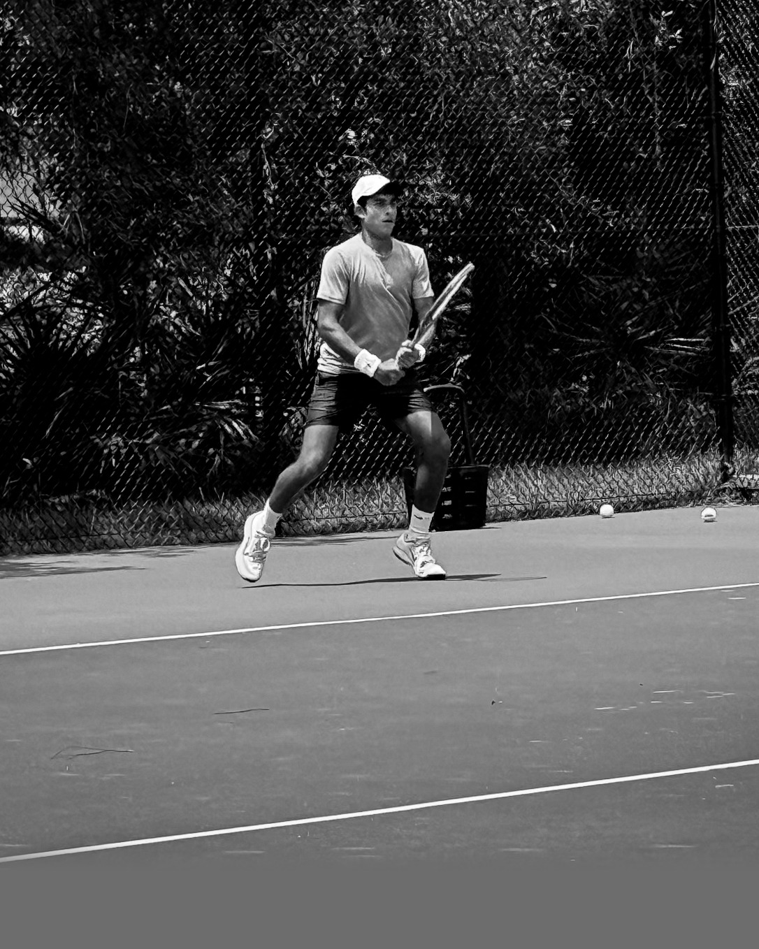 Shaan setting up for a two-handed backhand on a practice court, fence and palms behind